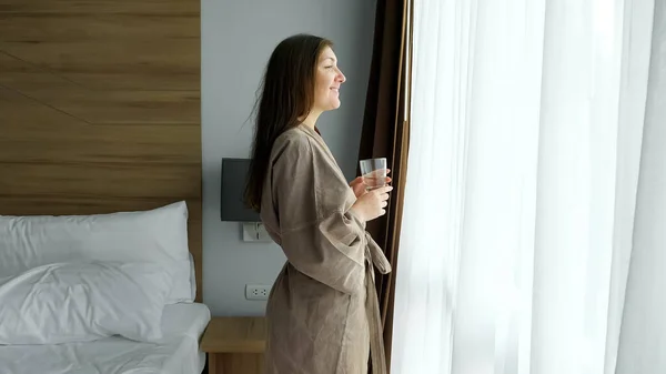 Mujer bebe agua limpia de pie cerca de la ventana en la habitación del hotel — Foto de Stock