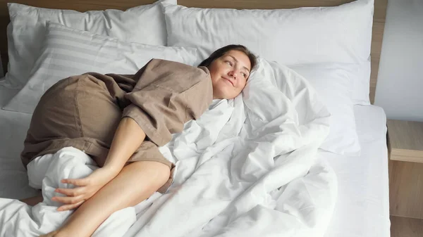 Mujer en albornoz descansa sobre una cama suave con manta y almohadas — Foto de Stock