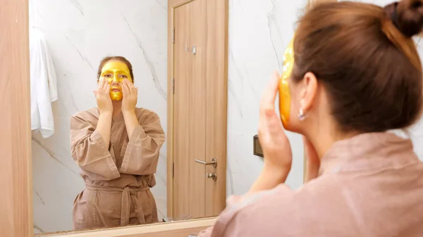 Frau mit goldener Maske massiert Gesicht im Hotelspiegel — Stockfoto