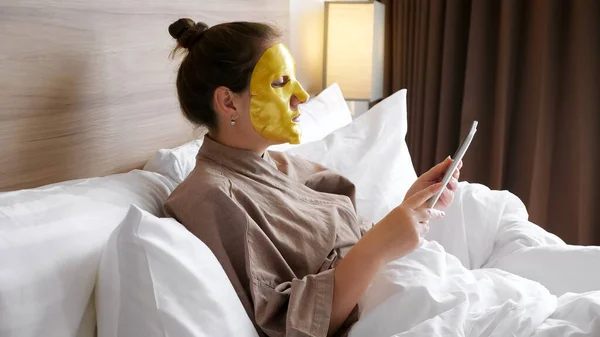 Senhora com máscara dourada surfa internet descansando em cama macia — Fotografia de Stock