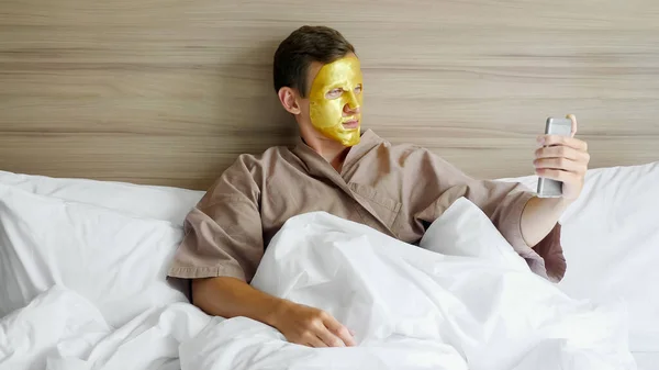 Парень в золотой маске делает селфи в мягкой постели в отеле — стоковое фото
