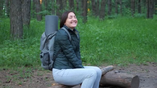 Ormanda bir kütüğün üzerinde sırt çantalı mutlu bir kadın oturuyor. — Stok video