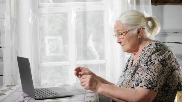 Mujer de pelo gris se pone gafas y tipos en la computadora portátil en la mesa sobre el fondo de la ventana — Vídeos de Stock