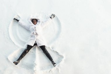 black girl lies on white forest glade and makes snow angles clipart