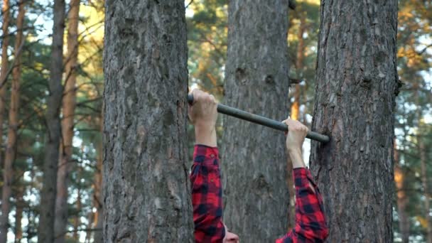 Man in een geruit shirt trekt zich nauwelijks aan een horizontale balk in het bos — Stockvideo