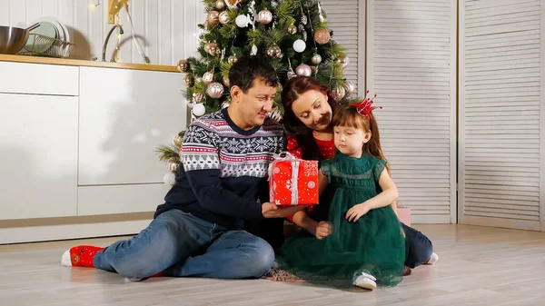 Menina criança descompacta caixa de presente com os pais pela árvore de Natal — Fotografia de Stock