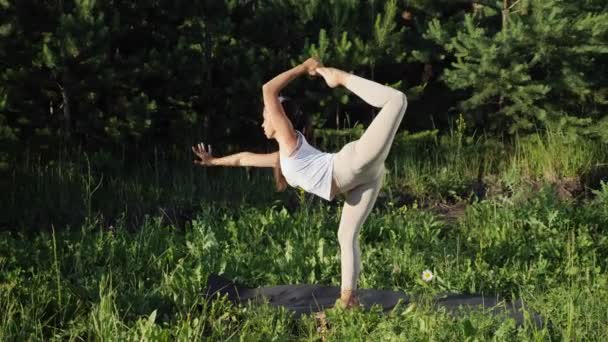 Mulher realiza asanas ioga, de pé em uma perna e um braço contra o fundo de pequenos pinheiros — Vídeo de Stock