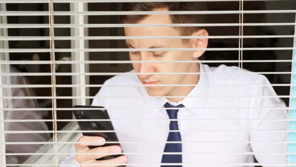 Man in white shirt and tie with phone, view through the blinds — Stock Video