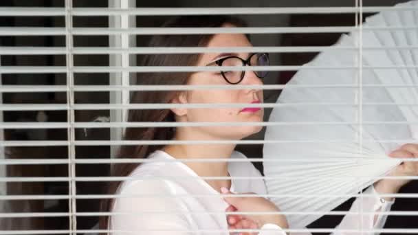 Vrouw met bril zit aan het raam en zwaait een ventilator, uitzicht door de jaloezieën. — Stockvideo