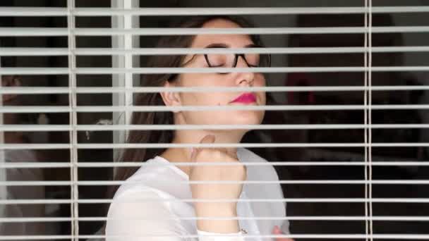Mladá žena sedí u okna a dívá se skrz žaluzie. Horké letní koncepce bez klimatizace — Stock video
