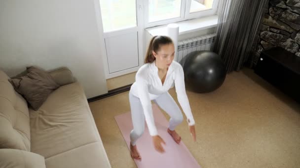 Meisje in wit trainingspak doet hurken op mat in licht kamer — Stockvideo