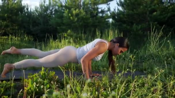 Красивая женщина йог выполняет упражнения на ковре на открытом воздухе на фоне маленьких сосен — стоковое видео