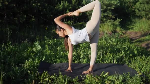 Kobieta wykonuje asany jogi, stojąc na jednej nodze i jednej ręce na tle małych sosen slow motion — Wideo stockowe