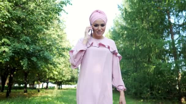 Belle femme dans un turban et une robe d'une couleur rose clair parlant au téléphone tout en marchant dans le parc — Video