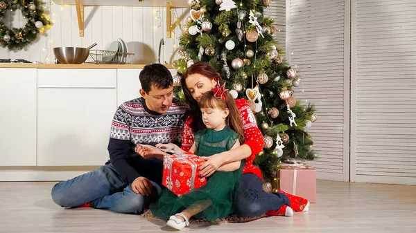 Menina criança descompacta caixa de presente com os pais pela árvore de Natal — Fotografia de Stock