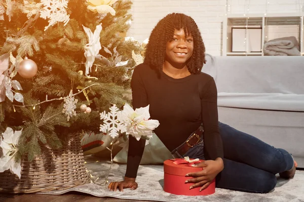 Leende svart flicka med lockigt hår sitter nära en julgran — Stockfoto
