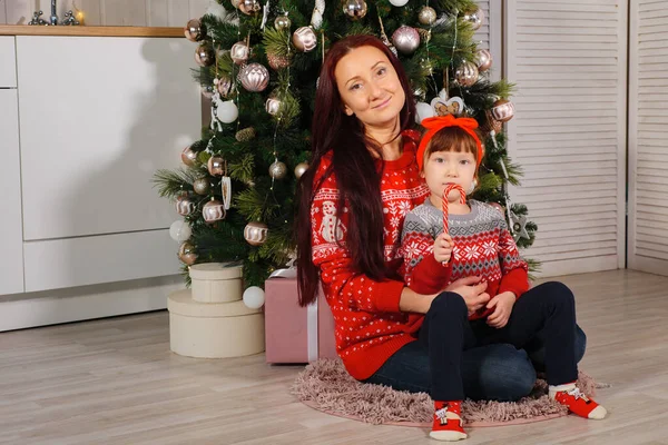 Η μαμά και η κόρη με τα κόκκινα πουλόβερ κάθονται στο χριστουγεννιάτικο δέντρο. — Φωτογραφία Αρχείου
