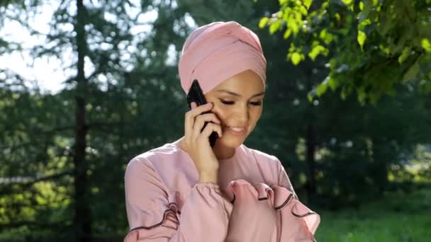 Jeune femme dans un beau turban et une robe de la même couleur rose douce parle au téléphone sur le fond d'arbres — Video