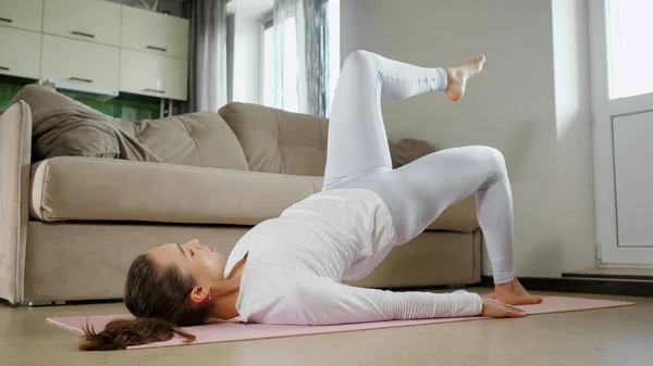 Woman does sports exercises lying on pink mat near couch — Stock Photo, Image