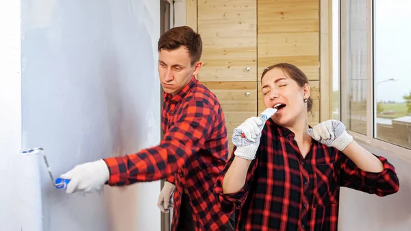 Žena zpívá a tančí, zatímco unavený muž maluje stěnu doma — Stock fotografie