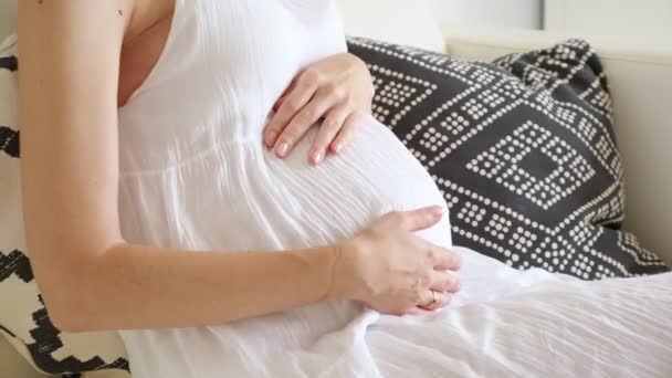 Female hands strokes belly sitting on sofa with soft pillows in light room at home — Stock Video