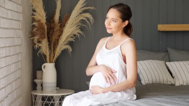 Pregnant lady in white dress strokes tummy sitting on bed — Stock Video