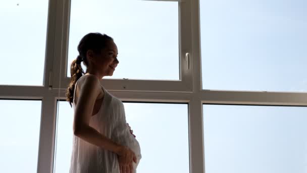 Pregnant woman in white dress strokes tummy near window — Stock Video