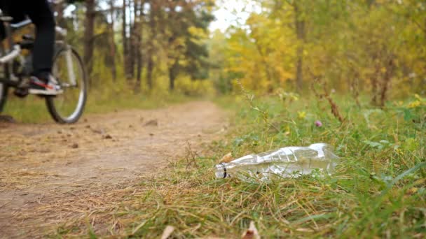 Primer plano de una botella de plástico en la hierba sobre el fondo de un ciclista en el bosque — Vídeo de stock