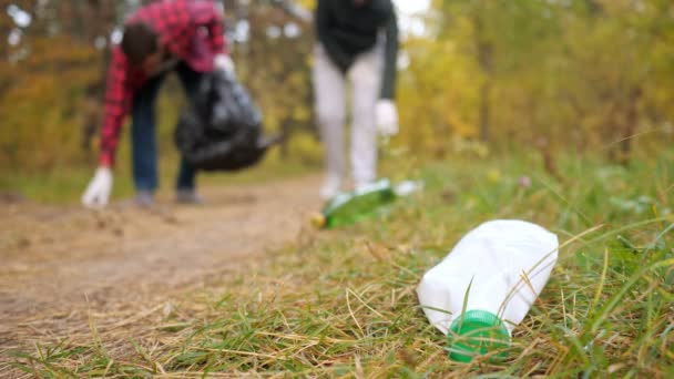 Primo piano di una bottiglia di plastica nell'erba sullo sfondo di una coppia che raccoglie spazzatura nella foresta — Video Stock
