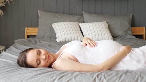 Donna incinta in abito bianco si trova sul letto con biancheria grigia — Video Stock