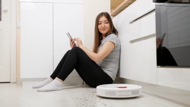 Vrouw met telefoon op de vloer robot stofzuiger verwijdert vuil — Stockvideo