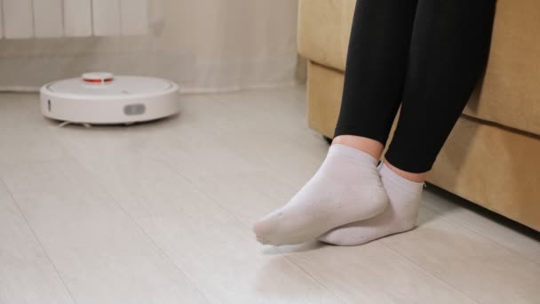 Perempuan dalam kaus kaki putih memungkinkan robot pembersih lulus duduk di sofa — Stok Video
