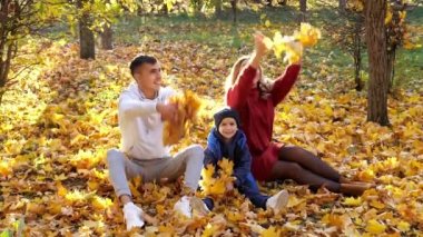 Güzel bir aile, güneşli sonbahar parkında kuru sarı yapraklar fırlatıyor.