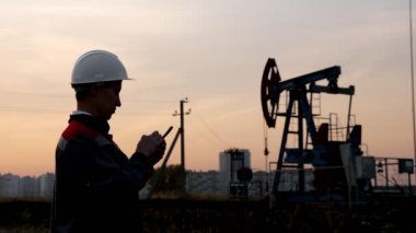 Beyaz miğferli, telefonlu bir adam sallanan bir petrol kuyusunun arka planında ve gün batımı gökyüzünde.