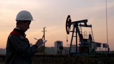 Tulumlu bir adam gün batımında bir petrol pompasının arka planına karşı bir kalemle kağıt dolduruyor.