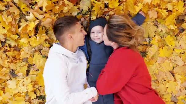 Madre y padre beso y abrazo chico acostado en hojas amarillas — Vídeo de stock
