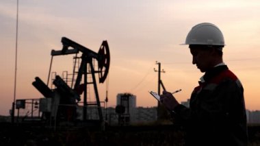 Beyaz kasklı bir adam gün batımında bir petrol pompasının arka planında bir kalemle kağıt dolduruyor.