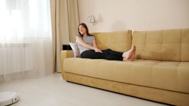 Kanepede uzanmış robot elektrikli süpürgenin çalışmalarını izleyen genç bir kadın.