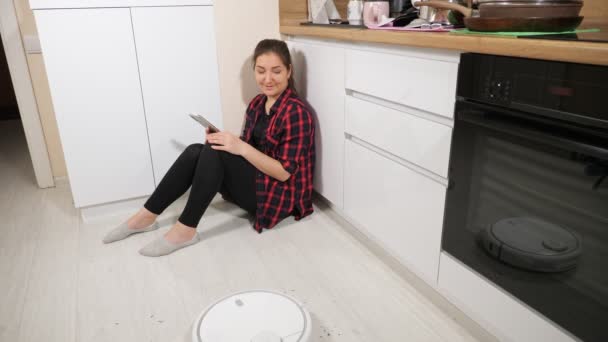 Mujer joven con el teléfono viendo robot con aspiradora sentado en el suelo de la cocina — Vídeo de stock
