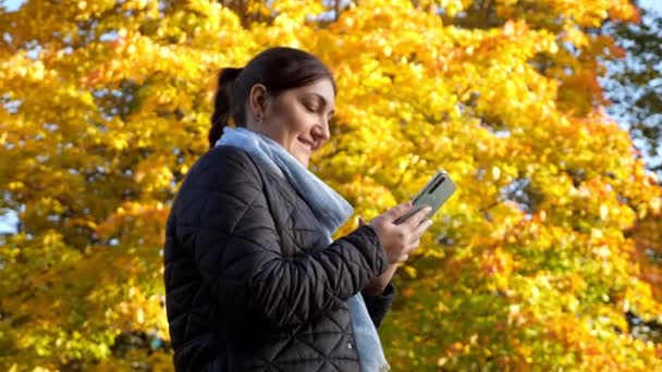 Giovane donna scrive un messaggio al telefono sullo sfondo di alberi gialli — Video Stock