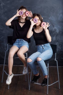 Sandalyelere siting ve having fun çörek ile oldukça mutlu genç kızlar. Siyah arka plan üzerine çörek ile neşeli gülümseyen kız portresi. İyi bir ruh hali, diyet kavramı.