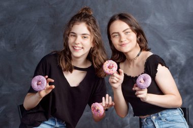 Sandalyelere siting ve having fun çörek ile oldukça mutlu genç kızlar. Siyah arka plan üzerine çörek ile neşeli gülümseyen kız portresi. İyi bir ruh hali, diyet kavramı.