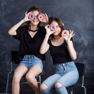 Sandalyelere siting ve having fun çörek ile oldukça mutlu genç kızlar. Siyah arka plan üzerine çörek ile neşeli gülümseyen kız portresi. İyi bir ruh hali, diyet kavramı.