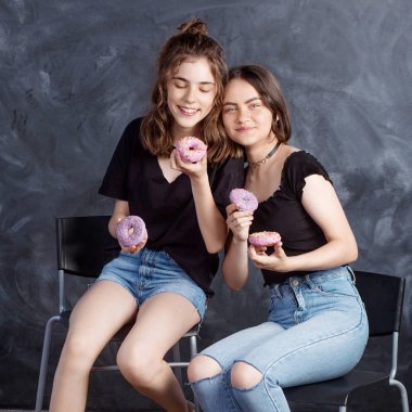Sandalyelere siting ve having fun çörek ile oldukça mutlu genç kızlar. Siyah arka plan üzerine çörek ile neşeli gülümseyen kız portresi. İyi bir ruh hali, diyet kavramı.