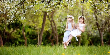 İki sevimli kız çiçek açması eski elma ağacı bahçede bir salıncak üzerinde eğleniyor. Güneşli bir gün. Çocuklar için bahar açık hava etkinlikleri