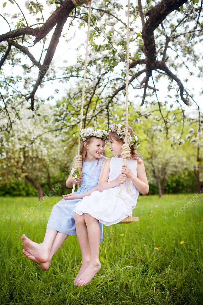 Two cute girls having fun on a swing in blossoming old apple tree garden. Sunny day. Spring outdoor activities for kids