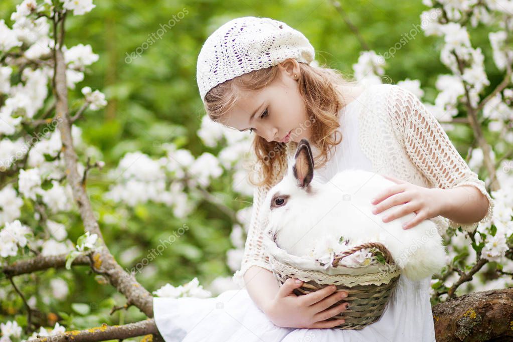 Beautiful  young girl in a white dress playing with white rabbit in the spring blossom garden. Spring fun activity for kids. Easter time