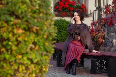 Şehir parkında bir bankta otururken akıllı telefon kullanan çekici bir orta sınıf kadın. Sonbahar geldi. Güzel bayan telefonda konuşuyor.