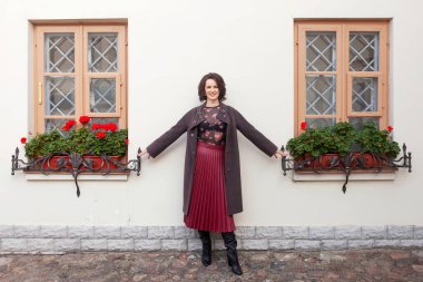 Güzel, gülümseyen orta yaşlı bir kadın güneşli bir sonbahar ya da bahar gününde sokağın kenarında kalıyor. Kahverengi ceketli bir model..