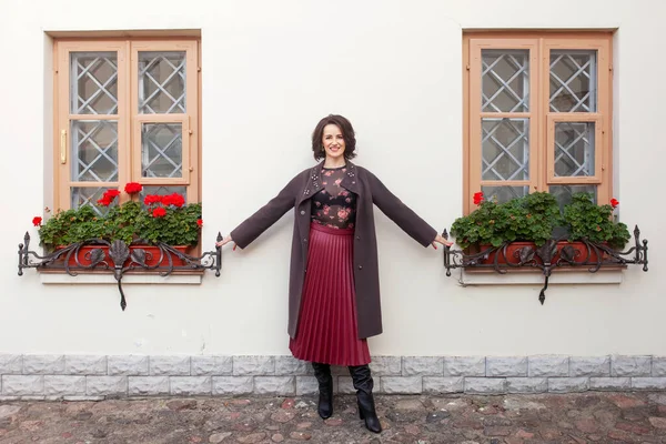 Güzel, gülümseyen orta yaşlı bir kadın güneşli bir sonbahar ya da bahar gününde sokağın kenarında kalıyor. Kahverengi ceketli bir model..